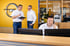 Drei Mitarbeiter im Opel-Autohaus beim Kundenservice am Telefon und bei einer Besprechung vor gelber Wand mit Markenlogo.