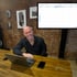 Ein lächelnder Mann sitzt an einem Holztisch mit einem Laptop und einem Telefon; im Hintergrund eine Ziegelwand und ein Kalender.