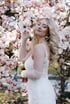 Blonde Frau im weißen Hochzeitskleid mit Spitzenhandschuhen posiert vor einem blühenden Magnolienbaum mit rosa Blüten.