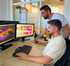 Zwei Männer arbeiten an zwei Computermonitoren mit farbigen 3D-Modellen und Plänen in einem Büro.