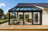 Modernes Glashaus mit dunklem Rahmen an einem Haus mit Holzterrasse und Garten.