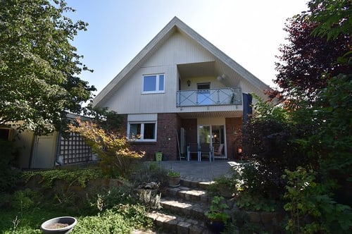 Haus mit Giebeldach, Balkon und Terrasse, umgeben von Bäumen und Sträuchern, bei Tageslicht.