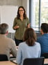 Lächelnde Referentin mit Brille hält einen Vortrag vor einer Gruppe in einem hellen Seminarraum mit Whiteboard.