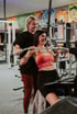 Two women exercising in a gym, one seated on a machine, the other standing behind her.