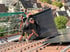 Arbeiter in Schwarz installiert schwarzes Solarpanel auf rotem Ziegeldach, andere Häuser im Hintergrund.