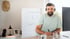 Lächelnder Mann mit Headset schreibt am Schreibtisch im Homeoffice, im Hintergrund ein Flipchart mit Notizen.