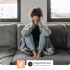 A woman with curly hair on a gray sofa, holding her head, looking downcast, for Chronic Fatigue.