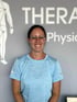 Lächelnde Physiotherapeutin mit Brille und blauem Shirt vor einer Wand mit der Aufschrift THERAPhysio.