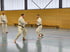 Zwei Männer in Karate-Anzügen üben Kampfsport in einer Sporthalle, ein dritter Mann steht im Hintergrund.