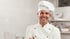Chef souriant en toque et veste de cuisine blanche, bras croisés, dans une cuisine lumineuse.