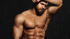 Shirtless muscular man with a beard posing with one arm raised against a dark background.