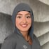 Junge Frau in grauer Uniform und Hijab lächelt, mit glattem grauen Hintergrund und welligem Muster rechts.
