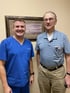 Two older men smiling, one in blue scrubs.

