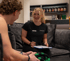 Lächelnde Fitness-Trainerin im Gespräch mit einem Kunden auf einem Sofa, während sie Notizen auf einem Klemmbrett macht.