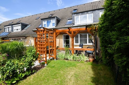 Hinterhof eines Reihenhauses mit Holzpergola, Gartenmöbeln, Rasen und grünen Pflanzen unter blauem Himmel.