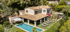 Eine große beige Villa mit Terrakotta-Dach, Pool, Rasen und Sonnenliegen umgeben von Bäumen.