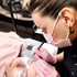 Wimpernstylistin mit rosafarbenen Handschuhen appliziert künstliche Wimpern auf die Augen einer liegenden Frau.