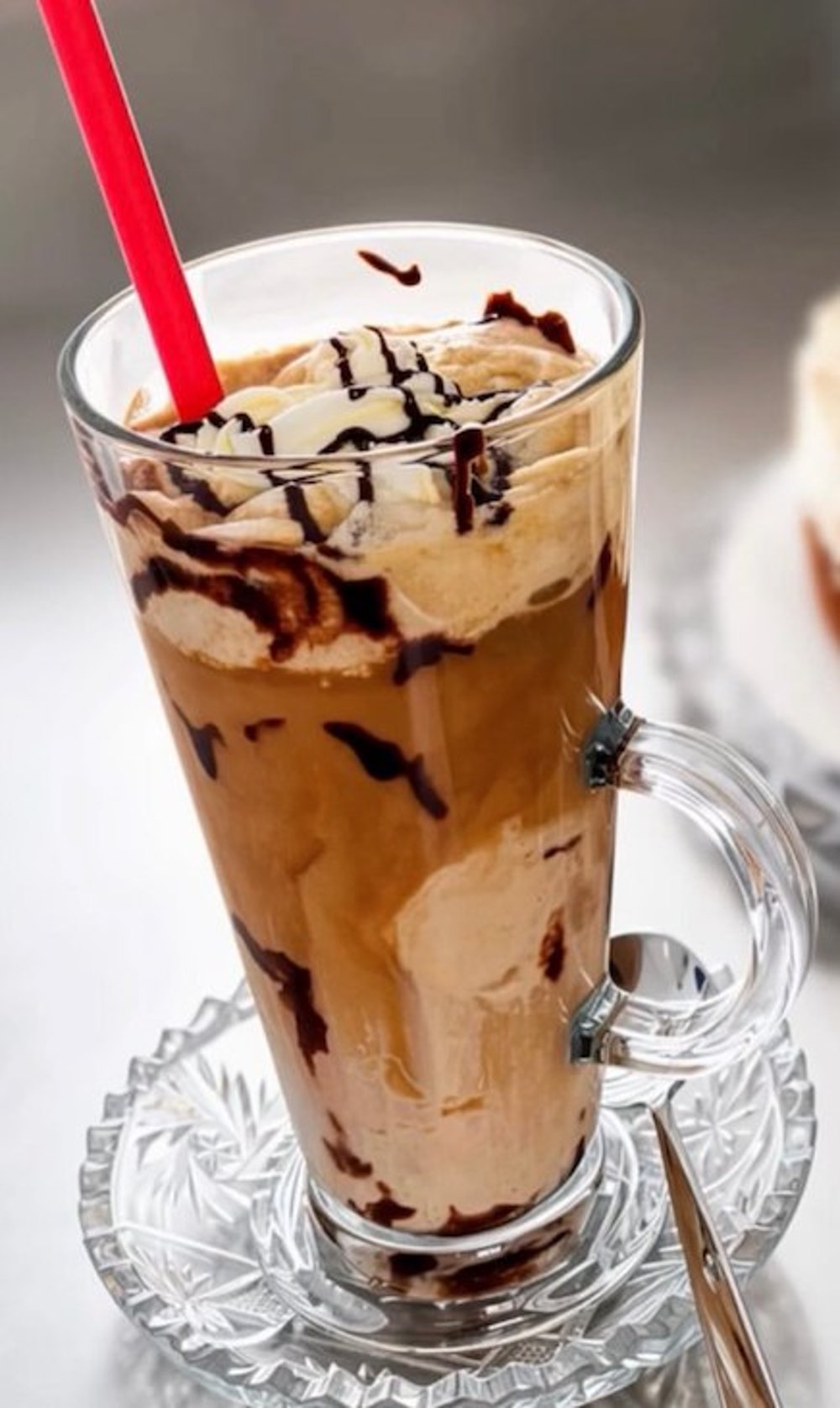 Eiskaffee im hohen Glas mit Sahne, Vanilleeis und Schokoladensauce, roter Strohhalm, auf einer Kristallglas-Untertasse