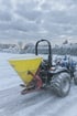 Blauer Traktor mit gelbem Streuer auf einer verschneiten Straße an einem bewölkten Wintertag.