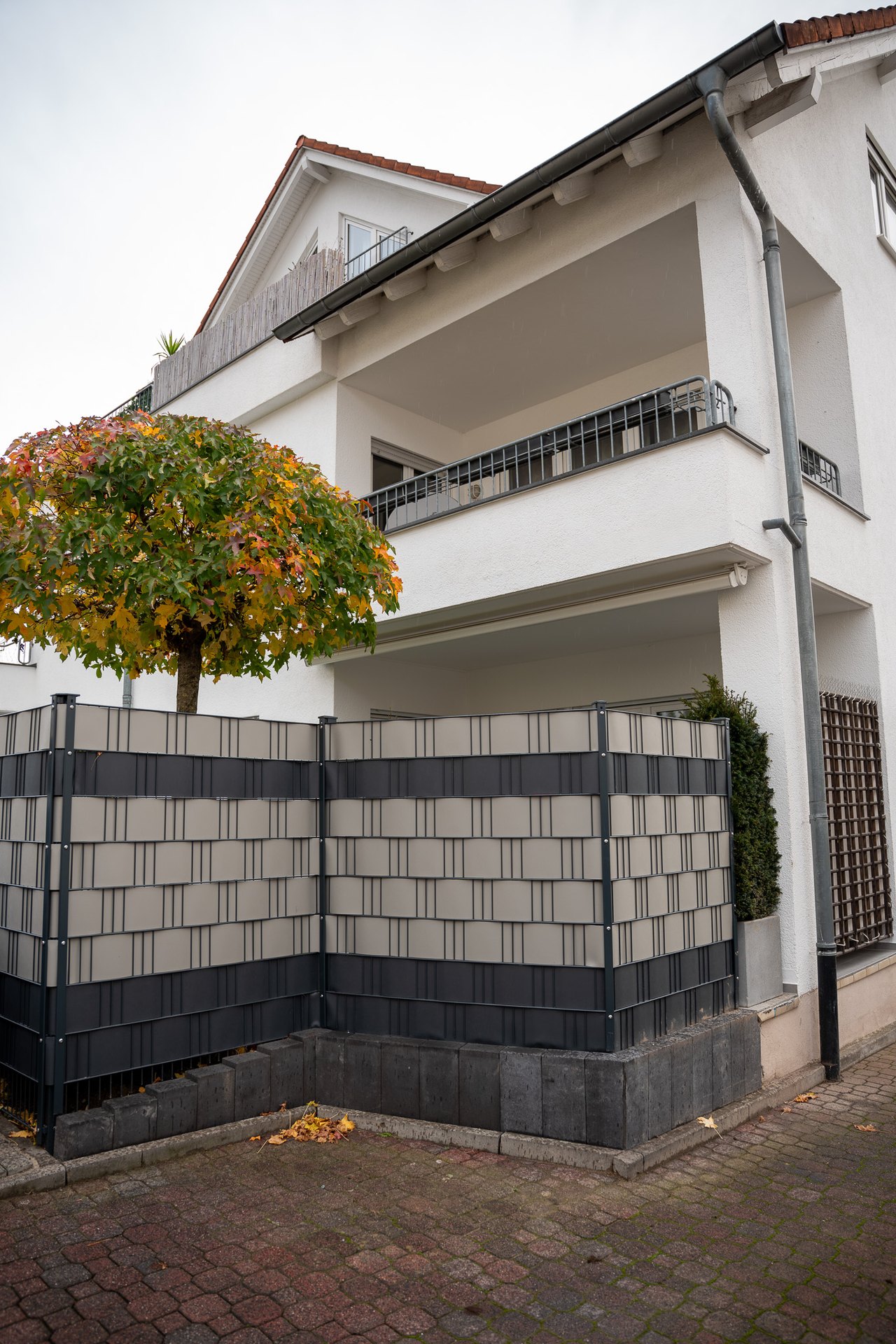 Weisses Haus mit rotem Dach, davor ein bunter Baum und ein hoher Sichtschutzzaun