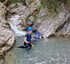 Zwei Personen in Neoprenanzügen und blauen Helmen stehen im knietiefen Wasser eines felsigen Bergbachs.