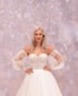 Blonde Frau in einem weißen Brautkleid mit Korsett-Oberteil und transparenten Puffärmeln vor rosa Bokeh-Hintergrund.