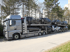 Silberner Autotransporter-LKW mit sieben dunklen PKWs auf zwei Ebenen unter blauem Himmel.