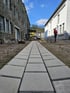 Ein frisch gepflasterter Gehweg aus grauen Betonsteinen führt zwischen einem Natursteinbau und einem weißen Gebäude hindurch.