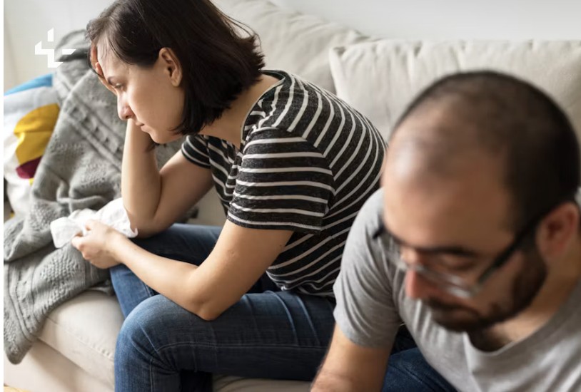 Eine Frau sitzt erschöpft auf dem Sofa, ihr Mann sitzt abgewandt daneben
