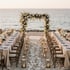 Strandhochzeit mit blumenverziertem Torbogen, zwei festlich gedeckten Tischen und Laternen am Meer.