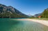 Ein türkisfarbener See mit hellem Sandstrand und bewaldeten Bergen unter blauem Himmel.