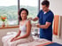 A male therapist in blue scrubs applies TENS unit electrode pads to a woman's shoulder in a clinical setting.