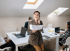 Smiling businesswoman in a brown suit talks on a phone, holding papers, in a brightly lit office with skylights.