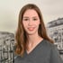 Junge Frau mit langen braunen Haaren und grauer Arbeitskleidung lächelt vor einer Schwarz-Weiß-Kulisse.