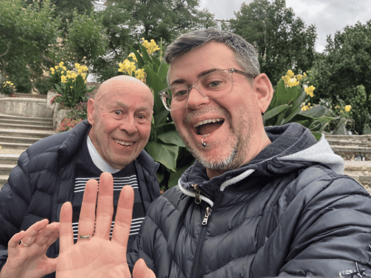 Two men smiling at the camera, one with glasses and a piercing waving, the other with a shaved head.