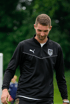 Ein junger Mann im schwarzen Trainingsanzug mit Puma-Logo und Vereinswappen blickt nach unten.