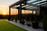 Moderne Glasveranda mit Schiebetüren und Möbeln, davor grüner Rasen bei Sonnenuntergang.