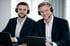 Zwei lächelnde Männer mit Headsets und Mikrofonen sitzen vor Laptops.