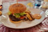 Ein großer Rindfleisch-Cheeseburger mit Sesambrötchen, Salat, Zwiebeln, Tomaten, Gurken und Pommes auf weißem Teller.