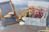 Miniatur-Liegestuhl neben einer Holzkiste voller Euro-Geldscheine auf Sand mit Muscheln und Seesternen.