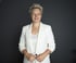 Frau mit kurzen blonden Haaren, weißem Blazer und weißem Oberteil lächelt vor dunklem Hintergrund.