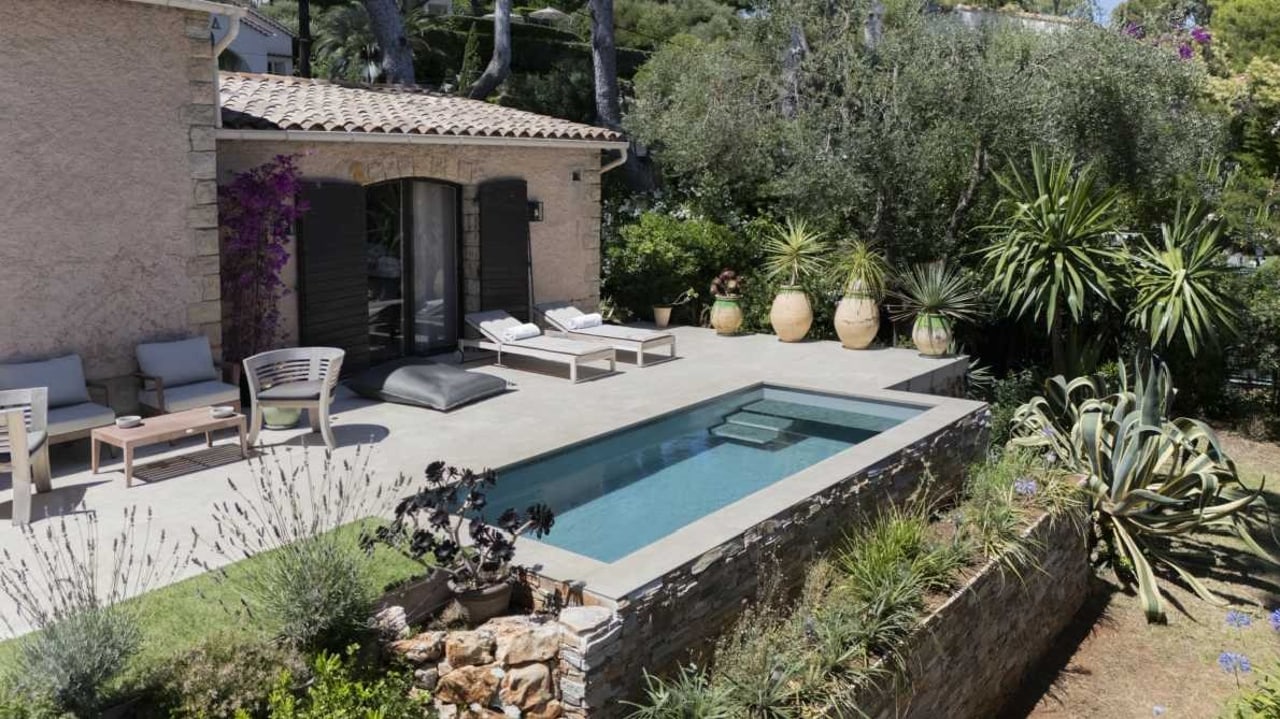 Rechteckiger Pool neben einem Haus mit Terrasse, Liegestühlen und Pflanzen im Garten.