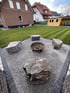 Eine rostige Feuerschale steht auf einer Kiesfläche, umringt von vier großen Sitzsteinen in einem gepflegten Garten.