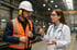Ein Bauarbeiter in Schutzkleidung spricht mit einer Ärztin mit Stethoskop in einer Fabrik.