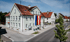 Helles Gebäude mit rotem Dach, Fahnen und der Aufschrift "RATHAUS" an einer Straße mit Bäumen und weiteren Häusern.