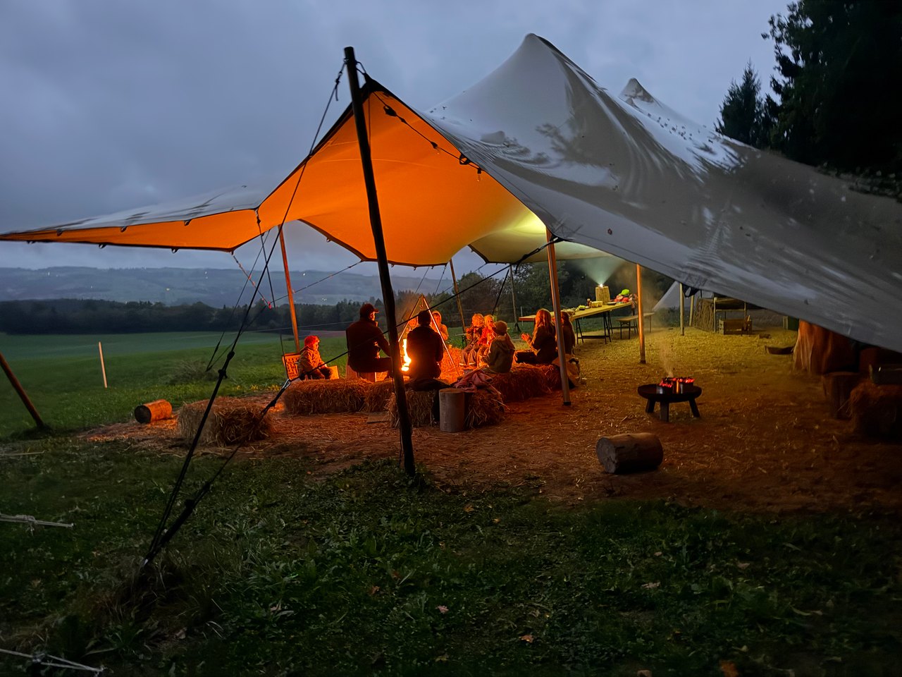 Village Hildisrieden: Feuerschale und Brennholz auf der Wiese, dahinter Sonnensegel, Holzbänke und ein Tipi mit Blick ins Hügelland