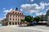 Historisches Rathaus am gepflasterten Marktplatz mit Arkaden, Uhrturm, grünen Bäumen und Außengastronomie unter blauem Himmel.