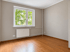 Leeres Zimmer mit Laminatboden, grauen Wänden, weißem Fenster, Heizkörper und Blick ins Grüne.