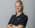 Lächelnde junge Frau mit blondem Haar im schwarzen Poloshirt mit grünem Logo vor grauem Hintergrund.