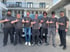 Sieben Männer in grauen T-Shirts mit orangefarbenem Logo stehen vor einem weißen Auto und zeigen aufeinander.